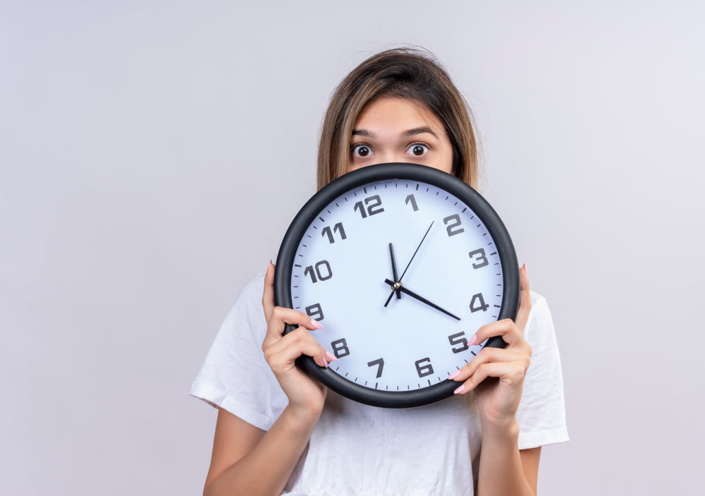 mujer-joven-sorprendida-camiseta-blanca-sosteniendo-reloj-pared-mientras-mira